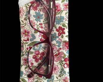 May include: A rectangular fabric pouch featuring a floral pattern with pink, red, and blue flowers on a cream background, secured with a white zipper at the top. A pair of dark red, tortoiseshell-patterned eyeglasses rests on the pouch surface.