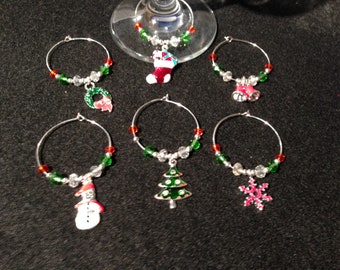 May include: A set of six silver-toned metal wine glass charms featuring holiday-themed pendants including a Christmas stocking, wreath, bells, snowman, tree, and snowflake, accented with red, green, and clear beads on circular wire hoops.