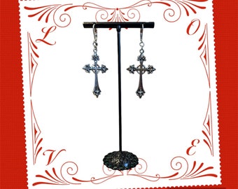 May include: Silver cross earrings with a detailed design. The earrings are hanging from a black stand against a white background.
