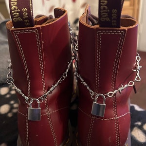 Puede incluir: Un par de botas de cuero burdeos con cordones negros y una cadena con un candado sujeto a cada bota. Las botas tienen una suela cosida amarilla y una etiqueta que dice "SOLES WITH BOUNCING"