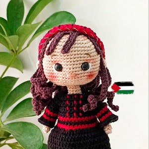 Könnte beinhalten: Häkelpuppe mit braunen Haaren, einem roten Stirnband und einem schwarzen Kleid mit roten Verzierungen. Die Puppe trägt ein traditionelles palästinensisches Kleid.