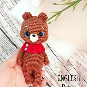 Könnte beinhalten: Ein brauner gehäkelter Teddybär mit blauen Augen und einem roten Schal. Der Bär steht auf einer Holzoberfläche. ENGLISH Pattern.
