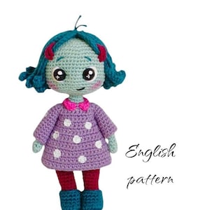 Puede incluir: Una muñeca de crochet con pelo turquesa, vestido morado con lunares blancos y pantalones rojos. La muñeca tiene un lazo rosa y ojos grandes. El texto "English pattern" está escrito debajo de la muñeca.