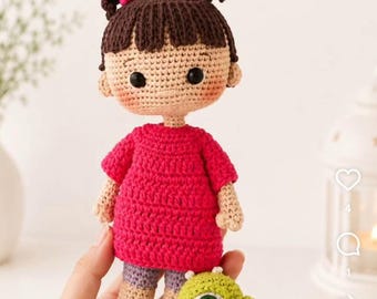 May include: A handmade crochet doll featuring a character with dark brown hair styled in two pigtails with pink hair ties, wearing a bright pink dress, light purple shorts, and white shoes, while holding a small green one-eyed monster toy.