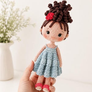 Pu&ograve; includere: Una bambola all'uncinetto fatta a mano con capelli ricci castano scuro raccolti in uno chignon alto con un piccolo fiore rosso. La bambola ha un incarnato color pesca chiaro, occhi neri e indossa un abito azzurro senza maniche con gonna a balze.