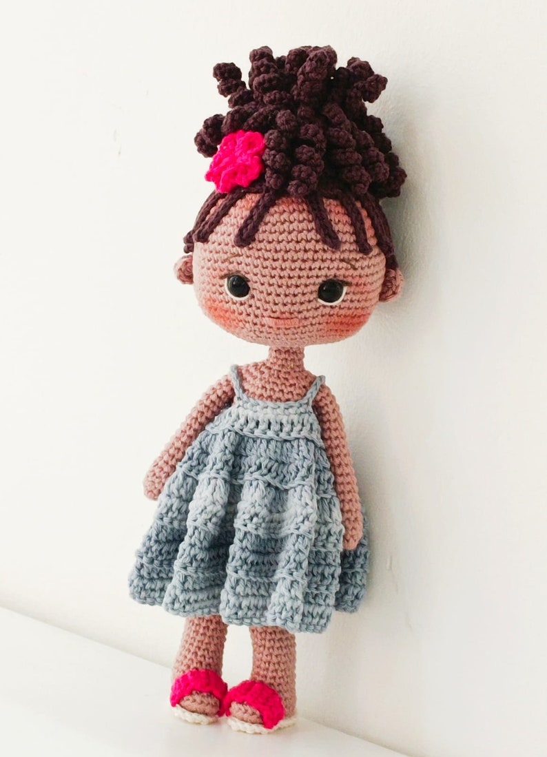 Pu&ograve; includere: Una bambola all'uncinetto fatta a mano con capelli ricci marrone scuro raccolti in uno chignon alto, decorata con un fiore rosa acceso. La bambola ha un incarnato marrone chiaro, guance rosate e indossa un vestito azzurro senza maniche.