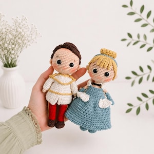 Puede incluir: Dos muñecos de ganchillo hechos a mano que representan una figura masculina con chaqueta blanca con ribetes dorados y pantalones rojos, y una figura femenina con vestido azul y moño amarillo. Ambos tienen ojos negros y mejillas rosadas.