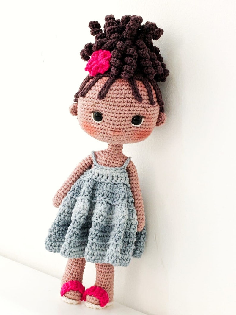 Pu&ograve; includere: Una bambola all'uncinetto fatta a mano con capelli ricci castano scuro raccolti in uno chignon alto, decorata con un fiore rosa acceso. La bambola ha occhi neri, guance rosate e indossa un vestito azzurro senza maniche con scarpe rosa.