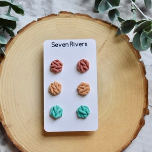 Könnte beinhalten: Ein Set mit drei Paaren sechseckiger Ohrstecker. Die Ohrringe sind aus Polymer Clay gefertigt und haben eine gestrickte Textur. Die Farben sind ein helles Pfirsich, ein dunkleres Pfirsich und ein helles Türkis.