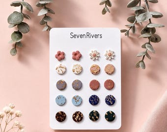 May include: A set of ten pairs of stud earrings displayed on a white card labeled Seven Rivers. The collection includes two pairs of flower-shaped studs and eight pairs of circular studs in various colors, including blue, pink, and dark green, with gold-toned flecks.