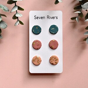May include: A white card displays six pairs of round stud earrings. The earrings are in three colors: teal, dusty rose, and peach. Each earring has gold leaf accents. The card reads "Seven Rivers" in black text.