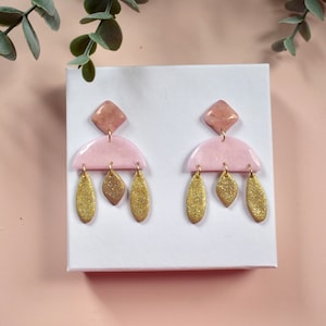 May include: A pair of pink and gold dangle earrings with a geometric design. The earrings feature a pink half-circle with a smaller pink diamond shape on top. Three gold leaf shapes dangle from the bottom of the half-circle.