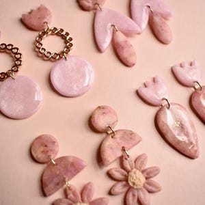 May include: A set of pink polymer clay earrings featuring a variety of shapes and designs, including flowers, circles, and abstract forms. The earrings are handmade and have a delicate, feminine aesthetic.