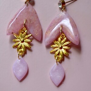 May include: A pair of pink and gold dangle earrings with a flower charm and a teardrop-shaped pendant. The earrings are made of resin and have a sparkly finish.