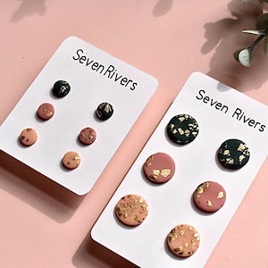 Op de afbeelding: Twee paar oorbellen op witte kaartjes met de tekst "Seven Rivers". De oorbellen zijn gemaakt van klei en hebben een gespikkeld ontwerp met gouden accenten. De ene set is roze en de andere is groen.