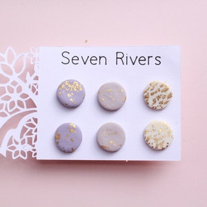 Peut inclure: Six paires de boucles d'oreilles rondes en céramique avec une base lavande et des paillettes dorées. Les boucles d'oreilles sont présentées sur une carte blanche avec le texte "Seven Rivers".
