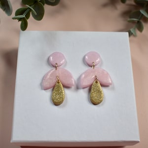 May include: A pair of handmade earrings. Each earring has a light pink, fan-shaped top and a gold, teardrop-shaped bottom. The earrings are displayed on a white surface, with green foliage in the background. The earrings are a stylish accessory.