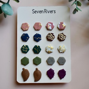 Puede incluir: Una tarjeta de exhibición con el texto "Seven Rivers" que muestra 12 pares de pendientes de botón hexagonales y en forma de hoja en varios colores y patrones, incluidos verde, azul, rosa, gris, marrón y dorado.