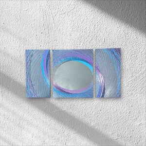 May include: A three-panel wall mirror with a silver oval center and swirling blue, purple, and silver designs. The panels are rectangular and mounted on a white textured wall. The design features curved lines.