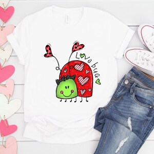 K&ouml;nnte beinhalten: Wei&szlig;es T-Shirt mit einem roten Marienk&auml;fer-Design und dem Text "Love Bug" in schwarzer und gr&uuml;ner Schrift.
