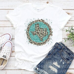 May include: White t-shirt with a turquoise and brown leopard print Texas state design. The design is surrounded by white paint splatters.