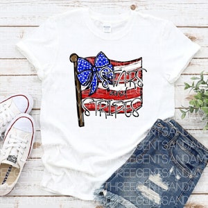 May include: White t-shirt with a red, white, and blue American flag design. The flag has a blue bow on top and the text "Stars and Stripes" written on it.