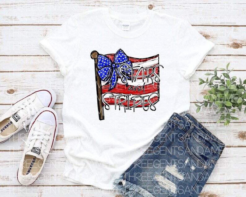 May include: White t-shirt with a red, white, and blue American flag design. The flag has a blue bow on top and the text "Stars and Stripes" written on it.