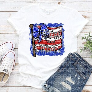 May include: White t-shirt with a red, white, and blue American flag design. The flag has a blue bow on the top and the text "Stars and Stripes" written on it.