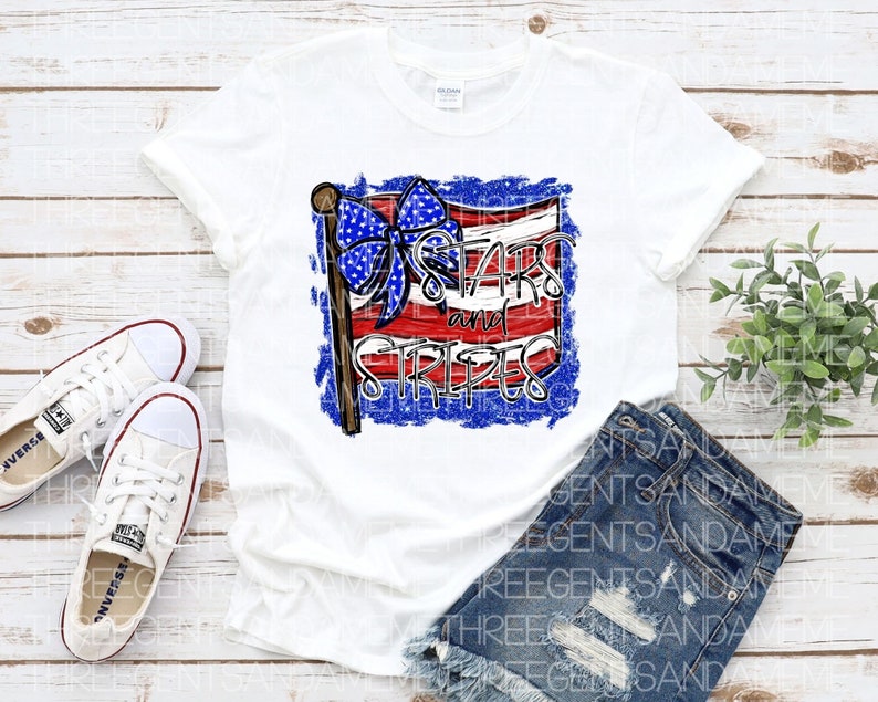 May include: White t-shirt with a red, white, and blue American flag design. The flag has a blue bow on the top and the text "Stars and Stripes" written on it.