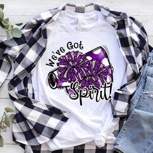 Op de afbeelding: Wit T-shirt met een paarse en witte megafoon met pompoenen en de tekst "We've Got Spirit!"