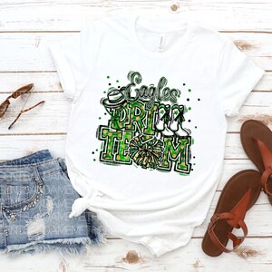 May include: White t-shirt featuring a green, black and gold graphic that reads "Eagles Cheer Team". The design includes a cheerleader's pom-pom, a hat and a pair of cheerleading shoes.