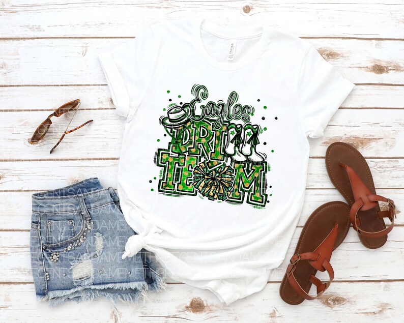 May include: White t-shirt featuring a green, black and gold graphic that reads "Eagles Cheer Team". The design includes a cheerleader's pom-pom, a hat and a pair of cheerleading shoes.