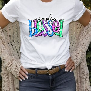 May include: A white t-shirt with the text "simply blessed" in colorful letters with polka dots and stripes.