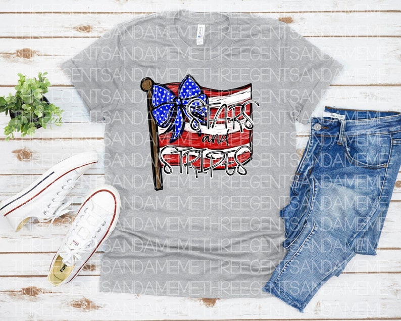 May include: Gray V-neck t-shirt with a red, white, and blue American flag design. The flag has a blue bow with white stars and the text "Stars and Stripes" written on it.
