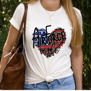May include: White t-shirt with a red, white, and blue heart design. The heart has the words "Air Force Wife" inside.
