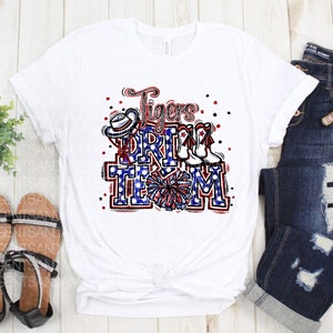 May include: White t-shirt featuring a red, white and blue graphic design that reads "Tigers Drill Team". The graphic includes a cheerleader, a hat and boots.