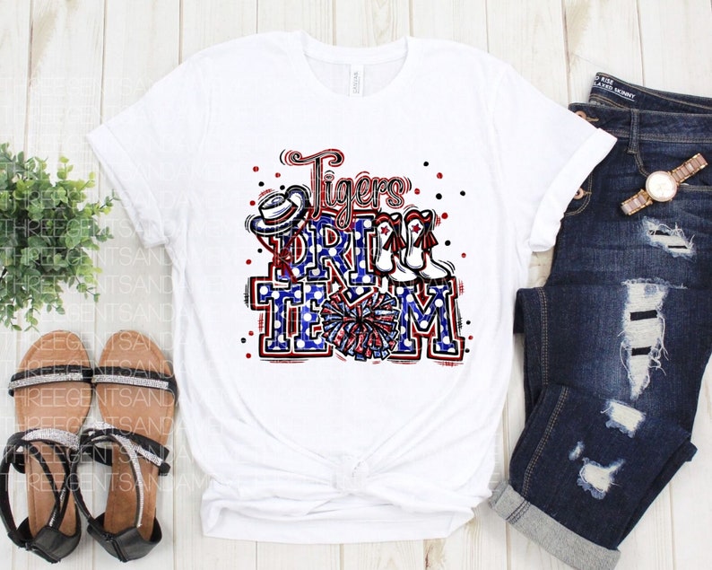 May include: White t-shirt featuring a red, white and blue graphic design that reads "Tigers Drill Team". The graphic includes a cheerleader, a hat and boots.