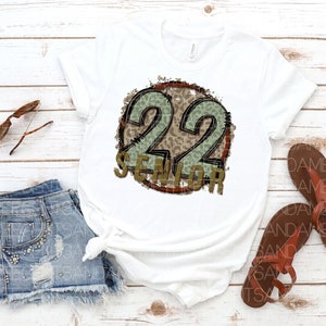 Puede incluir: Camiseta blanca con un gr&aacute;fico circular desgastado que presenta el n&uacute;mero 22 en estampado de leopardo verde y marr&oacute;n con la palabra "Senior" debajo.