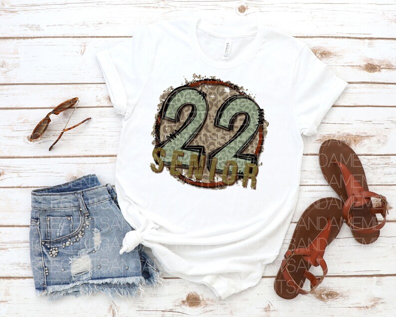 Puede incluir: Camiseta blanca con un gr&aacute;fico circular desgastado que presenta el n&uacute;mero 22 en estampado de leopardo verde y marr&oacute;n con la palabra "Senior" debajo.