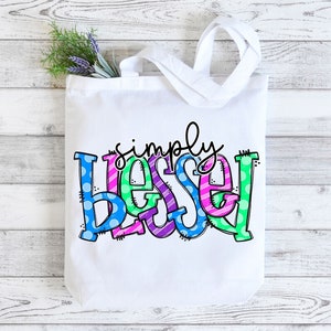 May include: White canvas tote bag with the words "simply blessed" in colorful, patterned letters. The letters are in shades of blue, green, pink, and purple.