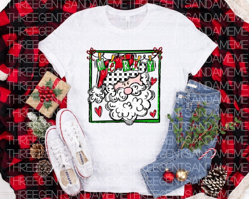May include: White t-shirt with a graphic of Santa Claus's face in a red and green frame. The frame has the word "Merry" written on it. The graphic is on a white background.