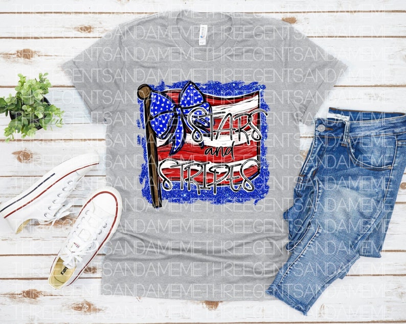 May include: A gray t-shirt with a distressed graphic of a red, white, and blue American flag with a blue bow. The text "Stars and Stripes" is written on the flag.