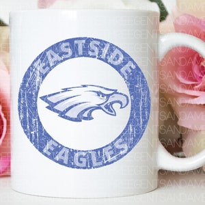 K&ouml;nnte beinhalten: Wei&szlig;e Keramiktasse mit einem blauen und wei&szlig;en, abgenutzten Kreisdesign. Der Kreis zeigt "Eastside Eagles" mit einer blauen Adlergrafik im Inneren.