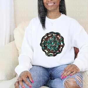 K&ouml;nnte beinhalten: Wei&szlig;er Pullover mit einem t&uuml;rkisfarbenen und braunen kreisf&ouml;rmigen Design mit einer stilisierten Blume.