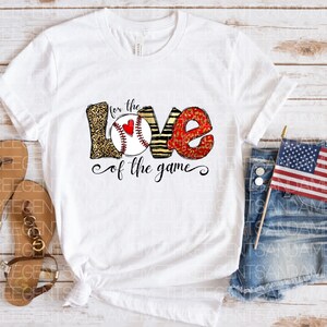 May include: White t-shirt with a graphic of the word "LOVE" with a baseball inside the "O". The letters are decorated with leopard print, stripes, and red glitter. The text "for the love of the game" is below the graphic.