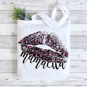 Pu&ograve; includere: Borsa tote bianca con un design a labbra con stampa leopardata nera e rosa e il testo "mamacita" in nero.