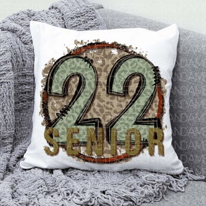 Puede incluir: Almohada blanca con un dise&ntilde;o circular marr&oacute;n y verde. El c&iacute;rculo tiene un patr&oacute;n de estampado de leopardo y el n&uacute;mero 22 en el centro. El texto "Senior" est&aacute; debajo del n&uacute;mero.