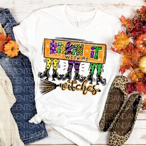 May include: White t-shirt with a colorful graphic design featuring three witches on broomsticks and the text "Kickin' it with my witches".