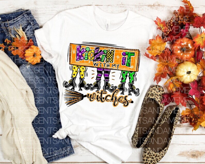 May include: White t-shirt with a colorful graphic design featuring three witches on broomsticks and the text "Kickin' it with my witches".