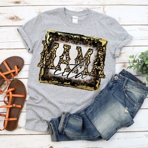 May include: Grey t-shirt with a gold and black square graphic featuring the text "Mamacita" in leopard print.